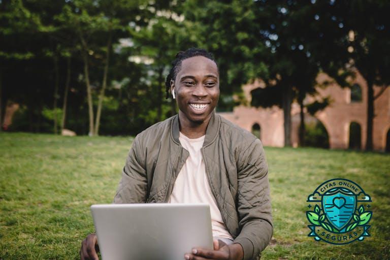 Positive African American male in casual clothes with wireless earbuds using laptop and smiling while sitting on green lawn and enjoying free time in quiet park