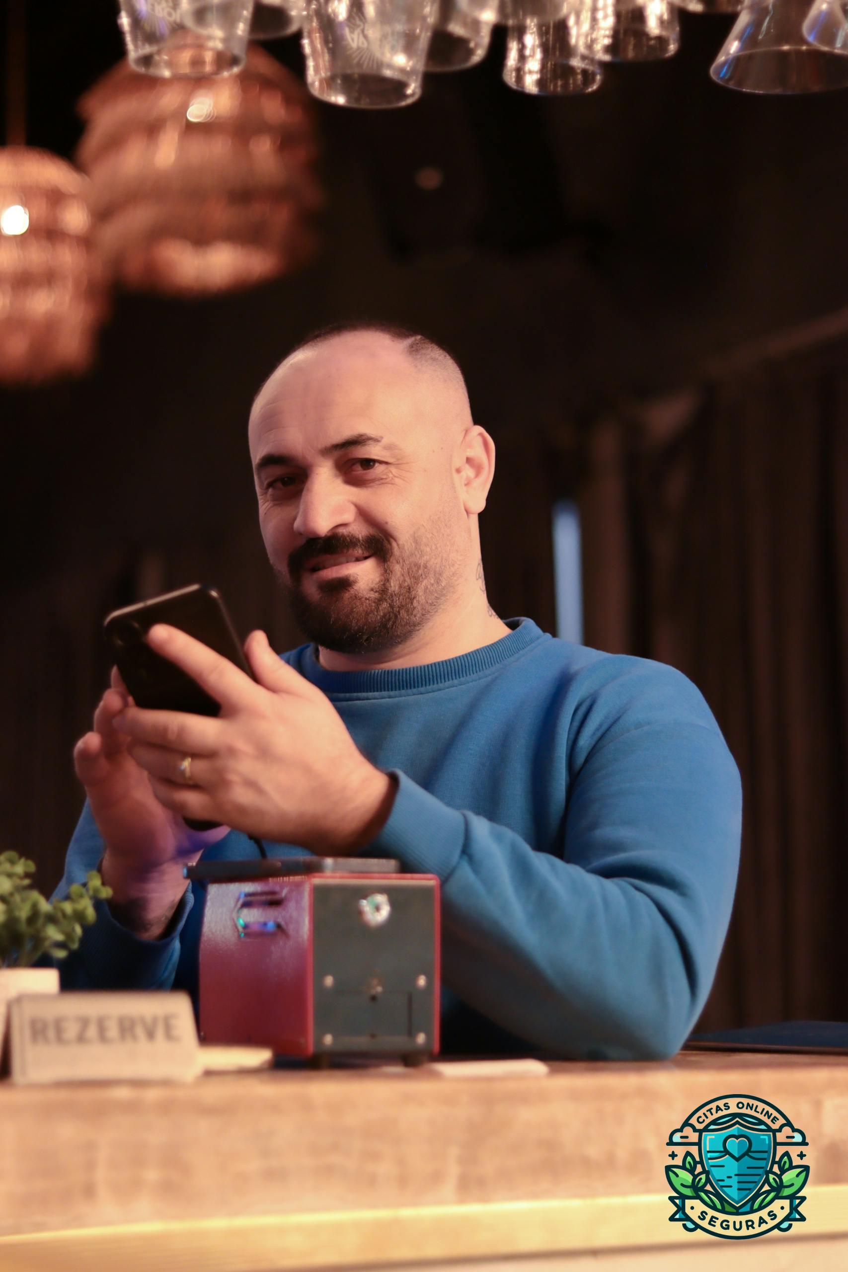 Smiling man in blue sweater using a smartphone, enjoying a relaxed moment in a cozy indoor setting.
